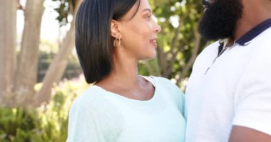 Çift ırklı kadın ve Afro-Amerikan erkek açık havada, yemyeşil bahçede birlikte gülümsüyorlar. Ağaçların arasından süzülen güneş ışığı, sıcak ve davetkar bir atmosfer yaratır, değişmez.