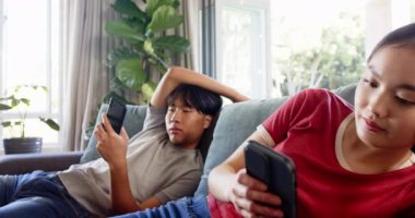 Kanepede dinlenmek, Asyalı kardeşlerin oturma odasında akıllı telefon kullanması. Teknoloji, yaşam tarzı, rahatlama, ev, bağlantı, iletişim