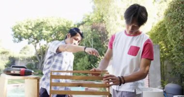 Asyalı baba ve genç oğul açık havada ahşap projede birlikte çalışıyorlar. Güneşli arka bahçeler yemyeşil ve etraflarını saran ağaçlar, huzur verici bir ortam yaratıyor, değişmeden.
