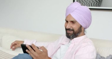 Sarıklı gülen adam evde kanepede dinlenirken tablet kullanıyor. Teknoloji, rahatlama, yaşam tarzı, dijital cihaz, ev içi, gündelik