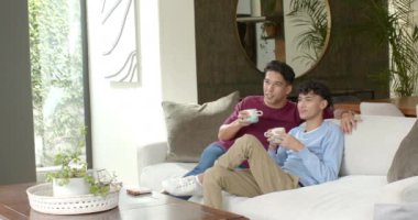 Kanepede dinlenmek, çok ırklı eşcinsel çift kahve içip evde televizyon izlemek. LGBT, rahatlama, konfor, arkadaşlık, bağlanma, rahatlama