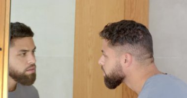 Sakallı, banyodaki aynaya bakan, görünüşüne odaklanan bir adam. Hijyen, kişisel bakım, tıraş, sabah rutini.