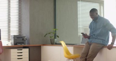 Akıllı telefon kullanarak, gülümseyen Afro-Amerikan bir adam modern ofis masasında oturuyor. Teknoloji, uzaktan çalışma, iş, serbest meslek, çalışma alanı, çağdaş, değiştirilmemiş, yaşam tarzı