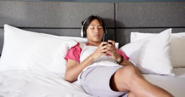 Akıllı telefon kullanarak, genç Asyalı adam yatakta oturuyor, ekrana odaklanıyor. Teknoloji, cep telefonu, kapalı alan, rahatlama, yatak odası, konsantrasyon