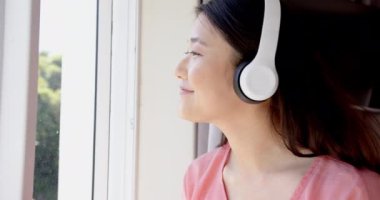 Müzik dinleyen, akıllı telefon kullanan ve pencere kenarında kulaklık takan bir kadın. Teknoloji, rahatlama, rahatlama, kapalı alan, modern