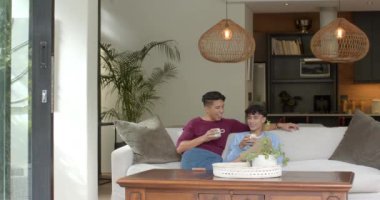 Evde dinlenen, çok ırklı eşcinsel çift kanepede oturup kahve içiyor. Rahatlama, ev hayatı, birliktelik, bağlanma, ilişki, rahatlık