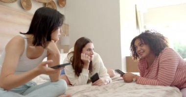 Sohbet ve gülüşmeler, üç kadın arkadaşın akıllı telefonlarıyla yatakta oturması. Arkadaşlık, kaynaşma, sosyal medya, teknoloji, rahatlama, rahatlama