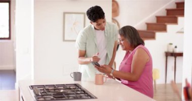 Son sınıf kadınına yardım etmek, mutfakta akıllı telefon özellikleri sergileyen genç bir adam. Yardım, teknoloji, yaşlılar, nesiller arası, iletişim, bakım