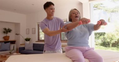 Oturma odasında gülümseyen erkek fizyoterapist, kol egzersizleri yapan yaşlı bir kadına yardım etmek. Fizyoterapi, rehabilitasyon, sağlık, yaşlılık, destek, terapi