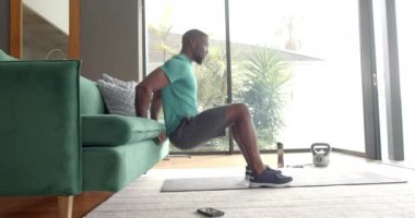 Güçlü ve formda bir adam evdeki koltuğu kullanarak triseps dalışı yapıyor, içeride egzersiz yapıyor. Fitness, egzersiz, güç, egzersiz, spor salonu, trisepsler, eğitim, değişmemiş, yaşam tarzı