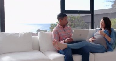 Hamile bir kadın, bir erkekle kanepede oturmuş laptopunu kullanıyor ve kahve içiyor. Aile, hamilelik, çift, teknoloji, rahatlama, birliktelik