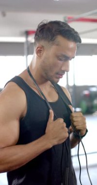 Dikey video: İp atlama, spor salonunda kaslı bir adam kameraya özgüvenle bakıyor. Fitness, atlet, egzersiz, antrenman, vücut geliştirme