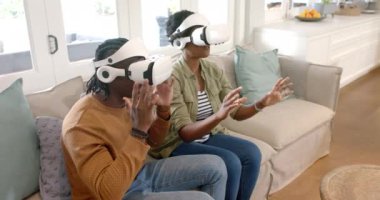 Afro-Amerikan çift, VR kulaklıklarını kullanarak koltukta oturup sanal dünyayla etkileşime geçiyorlar. Sanal gerçeklik, teknoloji, etkileşim, oturma odası, yenilik, eğlence