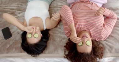 Evde dinlenirken, gözleri salatalık dilimleri ile yatakta uzanmış iki kadın arkadaş. Rahatlama, kendine bakma, şımartma, cilt bakımı, gençleşme, arkadaşlık.