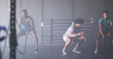 Spor salonunda savaş halatlarıyla antrenman yapan bir grup adam. Genç erkekler atletik kıyafetler giyiyorlar, yoğun spor yapıyorlar, güçlü ve formda fizikler gösteriyorlar, değişmemiş, yavaş çekim.
