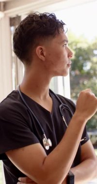 Dikey video: Steteskoplu erkek hemşire camdan dışarı bakıyor, hastane ortamını inceliyor. Sağlık, sağlık, medikal, tefekkür, sağlık hizmetleri uzmanı, hemşirelik, yansıma