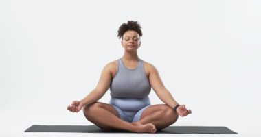 Büyük beden bir kadın yoga minderinde oturuyor, bir kolu başının üstünde uzanıyor. Açık mavi bir kıyafet giyiyor ve bir stüdyo ortamında değişmeden odaklanmış ve rahatlamış görünüyor.
