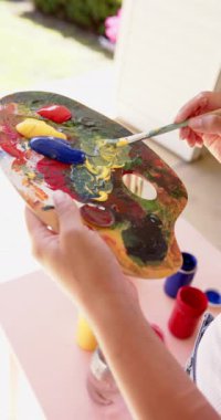 Dikey video: Palette canlı renkleri karıştırmak, sanatçı açık havada resim yapmaya hazırlanıyor. yaratıcılık, renkli, hazırlık, fırçalar, sehpa, ilham
