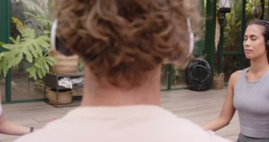 Yoga pozisyonunda meditasyon yapan, cam ev bahçesinde kulaklık takan bir kadın. Sağlık, farkındalık, rahatlama, meditasyon, teknoloji, doğa