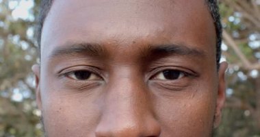 Afro-Amerikalı adam sıcak bir şekilde gülümsüyor, mutlu ve rahat görünüyor. Arka plan, yumuşak ışıklandırmalı ve rahat mobilyalı, değiştirilmemiş, yavaş çekimli rahat ev ortamını gösteriyor.