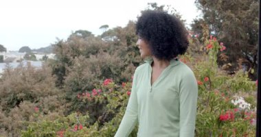 Yeşillik bahçesinin önünde canlı çiçeklerle gülümseyen melez kadın, kopyalama alanı. Ağaçlar ve uzak evler arka planda görülebilir, sakin bir açık hava ortamı yaratıyor, değişmemiş, yavaş çekim