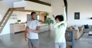 Modern oturma odasında birlikte yoga yapan mutlu genç çift. Arka planda mutfak, bitkiler ve rahat oturma alanı olan açık konsept alan, değiştirilmemiş, yavaş çekim