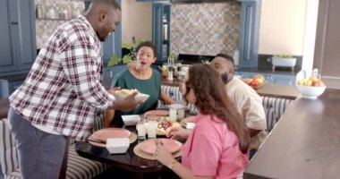 Yemek servisi yapan, Afro-Amerikan bir adam mutfaktaki yemek masasında çeşitli arkadaşlarıyla yemek yiyor. Arkadaşlık, toplanma, yemek, konukseverlik, yemek, iletişim.