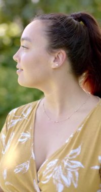 Dikey video: Açık havada gülümseyen kadın, günlük kıyafetlerle doğanın tadını çıkarıyor, halinden memnun görünüyor. Mutluluk, rahatlama, yaşam tarzı, keyif, yeşillik, rahatlık