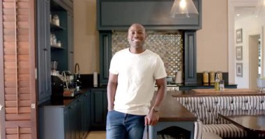 Gülümseyen Afrikalı Amerikalı adam modern mutfakta, günlük beyaz tişört ve kot pantolon giyiyor. Yaşam tarzı, ev, ev, mutluluk, iç mekan.