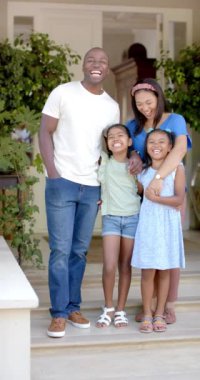 Dikey video: Dört kişilik gülümseyen aile evinin verandasında bir arada duruyor. Beraberlik, ön bahçe, açık hava, ebeveynler, çocuklar, mutluluk