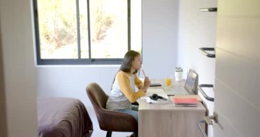 Çift ırklı genç bir kadın masada oturuyor, dizüstü bilgisayara bakıyor. Uzun kahverengi saçları, açık tenli, ve günlük kıyafetler giyiyor, değişmemiş, ağır çekimde.