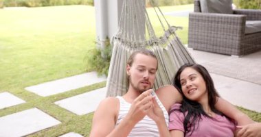 Bahçedeki hamakta dinlenen, akıllı telefon gösteren, fotokopi çeken bir çift. O, açık tenli, sakallı, kahverengi saçlı, telefonu tutan, esmer tenli, siyah saçlı, ekrana bakan.