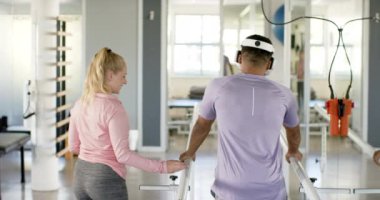 Protez bacaklı bir adam paralel barlarda terapistli VR kulaklık kullanıyor. Rehabilitasyon, fizyoterapi, sağlık, tıbbi, iyileşme, teknoloji, değiştirilmemiş, yavaş çekim