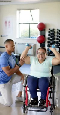 Dikey video: Tekerlekli sandalyedeki kadın spor salonunda yardımcı antrenörüyle dambılları kaldırıyor. Fitness, Egzersiz, Fizyoterapi, Sağlık, Sağlık, Ağır çekim