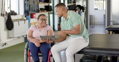 Engelli rehabilitasyonunda, tekerlekli sandalyede felçli bir kadına tablet gösteren bir adam, ikisi de gülümsüyor. Sağlık, rehabilitasyon, destek, terapi, takım çalışması, hasta, değişmemiş, yavaş çekim