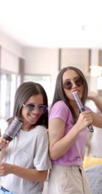 Dikey video: Şarkı söylemek ve eğlenmek, iki genç kız saç fırçalarını mikrofon olarak kullanıyor. Müzik, arkadaşlık, neşe, karaoke, neşe, gençlik
