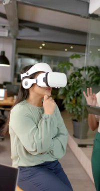Dikey video: VR vr kulaklık kullanan kadın, görev yerinde elinde tablet bulunan meslektaşıyla etkileşimde bulunuyor. Teknoloji, birlikte çalışma, işbirliği, sanal gerçeklik, iş dünyası, yenilik, yavaş çekim