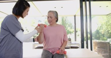Dambılları kaldıran, yaşlı Asyalı kadın ev terapisi sırasında fizyoterapistten yardım alıyor. Egzersiz, fitness, kuvvet eğitimi, rehabilitasyon, fizyoterapi, sağlık hizmetleri