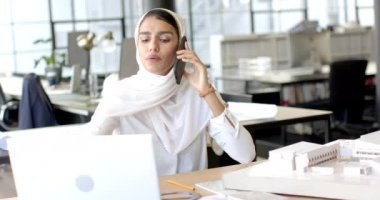 Akıllı telefondan konuşan, tesettürlü bir kadın modern ofiste dizüstü bilgisayarla çalışıyor. İş, teknoloji, iletişim, üretkenlik, çoklu görev, teknoloji meraklısı