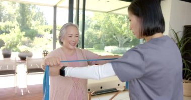 Kıdemli Asyalı kadın direniş bandıyla egzersiz yapıyor, evde fizyoterapistin yardımıyla. Egzersiz, fizyoterapi, sağlık, rehabilitasyon, fitness, terapi