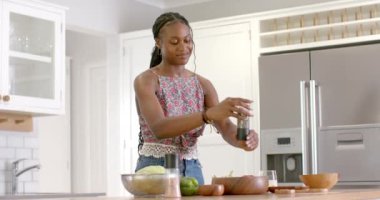 Çiçekli bluz giyen Afro-Amerikalı genç kadın evde, mutfakta yemek hazırlıyor. Saçlarını ördü, boncuklu bir kolyesi var ve biber öğütücü kullanıyor, değiştirilmemiş, yavaş çekim..