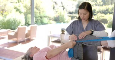 Fizyoterapist evde direniş bandı egzersizinde kıdemli Asyalı kadına yardım ediyor. Fizyoterapist, rehabilitasyon, yaşlılar, sağlık hizmetleri, fitness, terapi