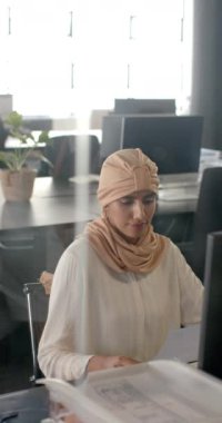 Dikey video: Ofiste çalışan, elinde belge tutan ve tesettürlü bir kadın. İş, meslek, şirket, çeşitlilik, güven, işyeri