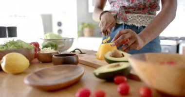 Genç bir Afro-Amerikan kadın evde, mutfakta sarı biber dilimliyor. Koyu kıvırcık saçları var, çiçekli önlük giyiyor ve etrafı taze sebzelerle çevrili.