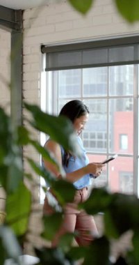 Dikey video: Tablet tutmak, iş kadını pencerenin yanında durup ofisin dışına bakmak. profesyonel, teknoloji, çalışma alanı, modern, yönetici, yavaş çekim