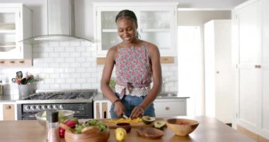 Genç bir Afrikalı Amerikalı kadın mutfakta, evde yemek hazırlıyor. Koyu renk örgülü saçları, çiçekli bluzu ve kot şortu var.