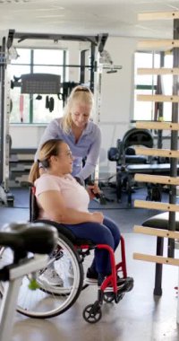 Dikey video: Dambılla egzersiz, tekerlekli sandalyedeki kadın spor salonunda terapistin yardımıyla. Spor, sakatlık, güç, eğitim, sağlık, egzersiz