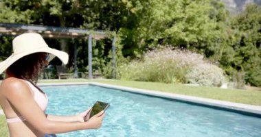 Havuz başında dinlenen, güneş şapkalı bir kadın tablet kullanıyor, yaz gününün tadını çıkarıyor, fotokopi çekiyor. Tatil, tatil, rahatlama, teknoloji, güneş banyosu, açık hava