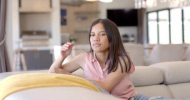 Evdeki kanepede oturan melez genç bir kadın, düşünceli görünüyor. Uzun kahverengi saçları, açık kahverengi teni var, pembe bluzu var, değişmemiş, yavaş çekim