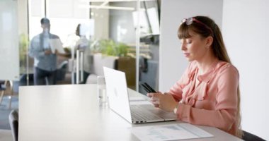 Akıllı telefon ve dizüstü bilgisayar kullanan kadın, modern ofiste çeşitli iş arkadaşlarıyla birlikte çalışıyor. İş, takım çalışması, işbirliği, teknoloji, üretkenlik, kurumsal